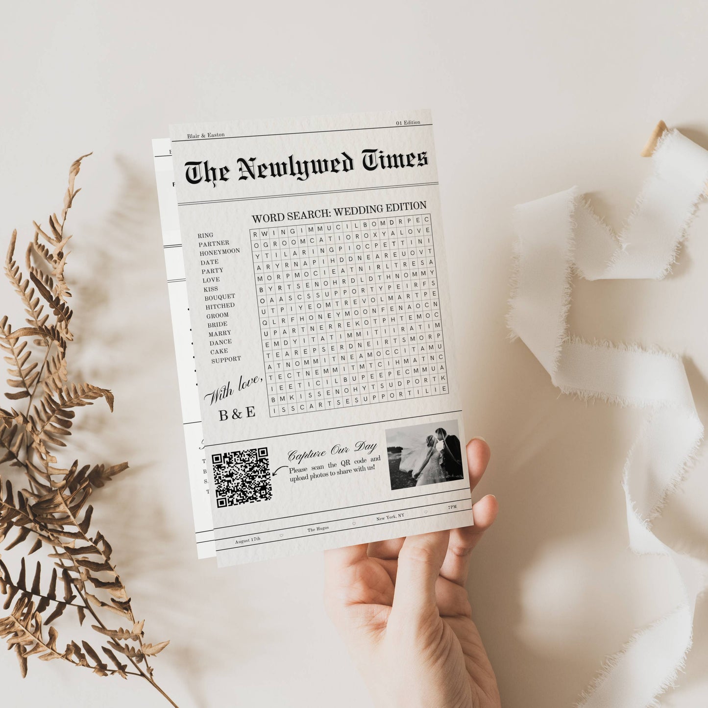Newspaper titled 'The Newlywed Times' held by a hand on a light background with dried leaves.