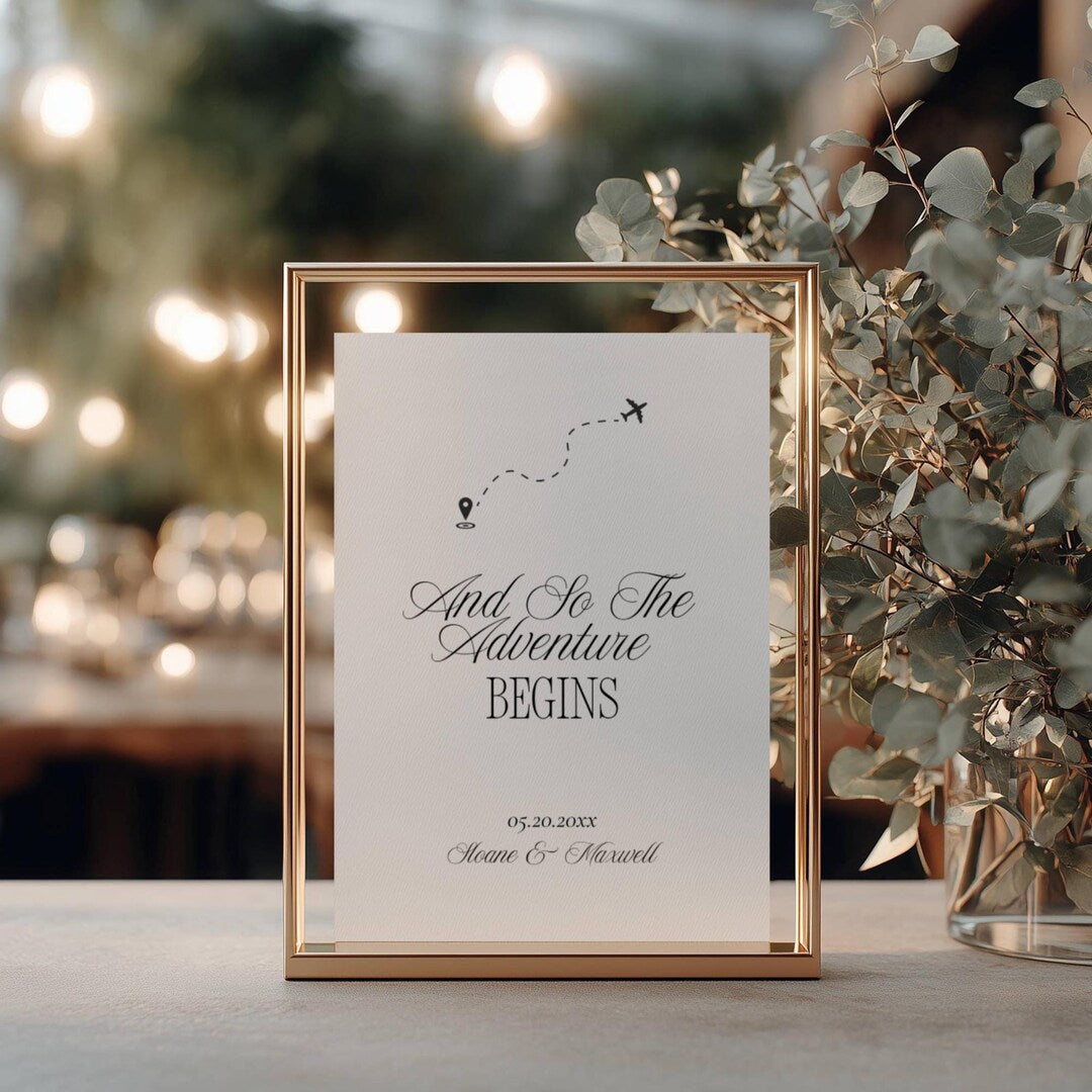 And So The Adventure Begins wedding sign in a gold metal frame with an airplane icon and dotted flight path, styled outdoors