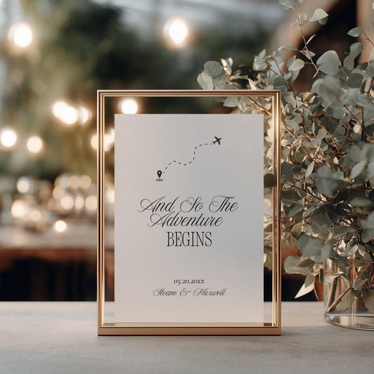 And So The Adventure Begins wedding sign in a gold metal frame with an airplane icon and dotted flight path, styled outdoors