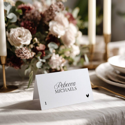 Old Money wedding place card with elegant script name and a hand-drawn rooster meal icon, styled on a white tablecloth with soft floral in the background