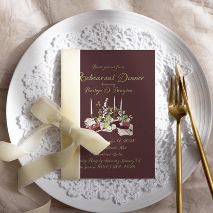 Burgundy rehearsal invitation styled with cream ribbon, candle and floral illustration, on a lace doily with gold forks