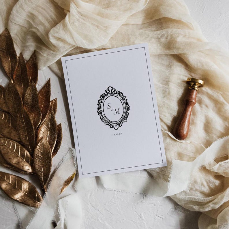 Wedding invitation with ornate black monogram in oval frame styled with gold leaf garland