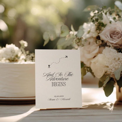 And So The Adventure Begins wedding sign flat lay showing black serif and script typography with an airplane and location pin icon