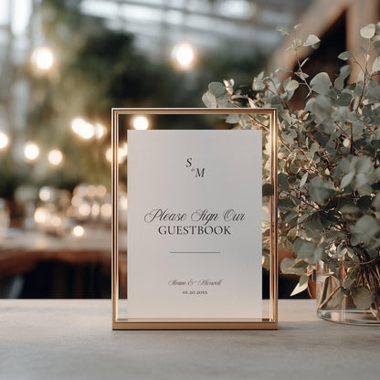 Old Money monogram wedding guestbook sign in a gold frame with cursive script, styled among soft foliage