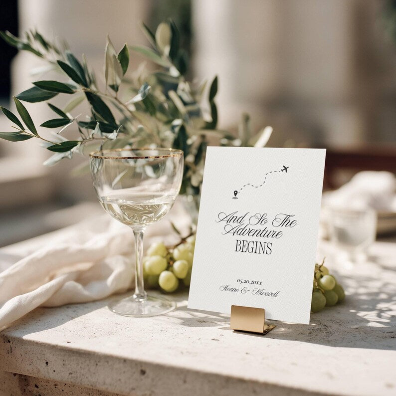 And So The Adventure Begins wedding sign displayed on a gold metal card stand