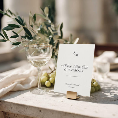 Old Money monogram wedding guestbook sign held by a gold clip at a reception table setting with olive branches