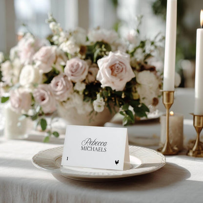 Reception table setting with a striped white plate, folded Old Money place card featuring a rooster meal icon, pink rose florals, and gold candlesticks