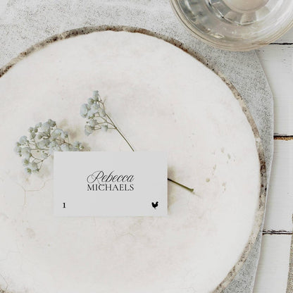 Old Money place card with elegant script name and hand-drawn chicken meal icon, resting on a textured plate with light blue florals