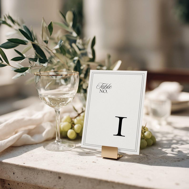 Minimalist wedding table number card with a black double-line border and Roman numeral I, displayed on a gold metal stand