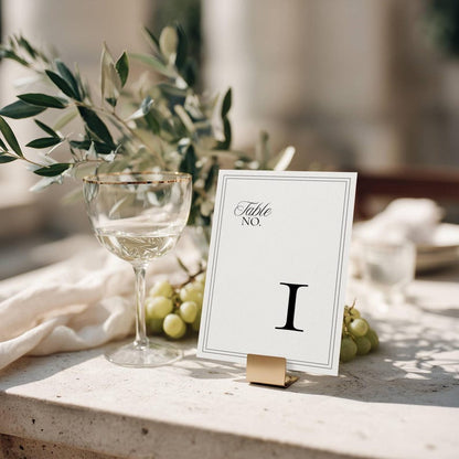 Minimalist wedding table number card with a black double-line border and Roman numeral I, displayed on a gold metal stand