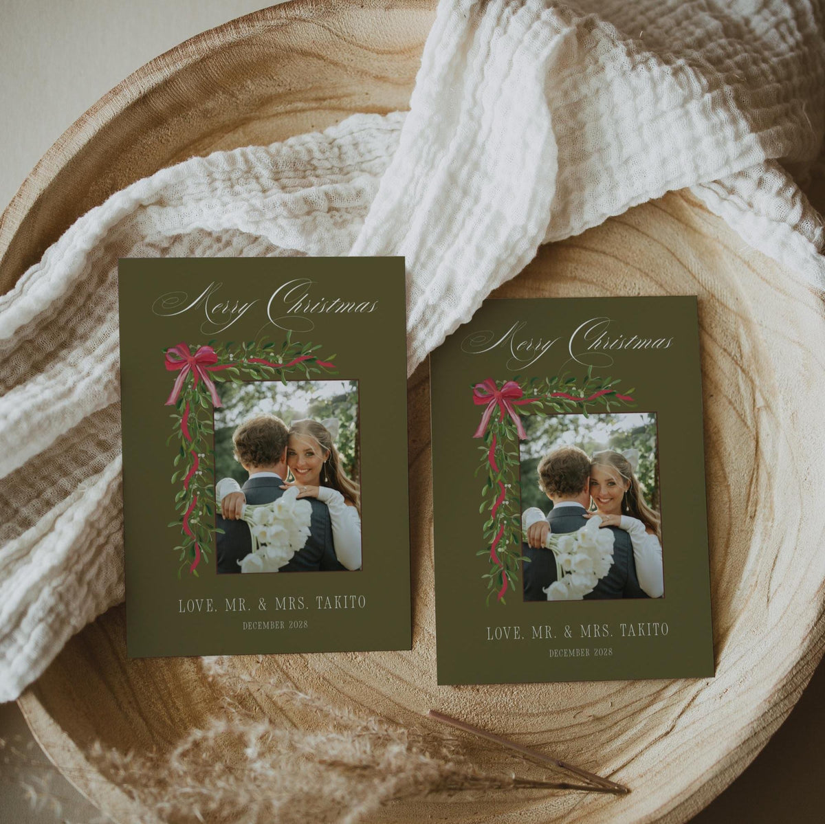 Pair of 5x7 olive green mistletoe newlywed holiday cards in a round wooden bowl with cream muslin