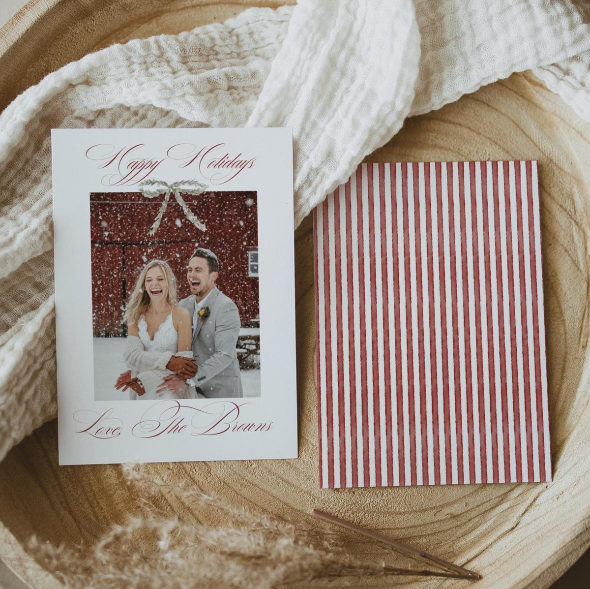 Happy Holidays newlywed Christmas photo card in deep red script beside its red striped back on a wooden board with cream gauze
