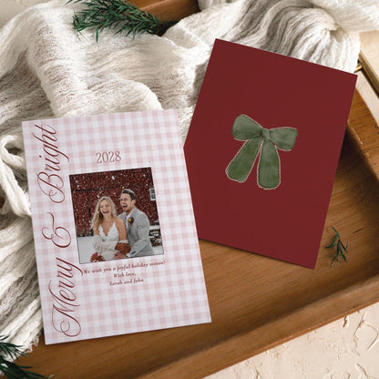 Red gingham Merry and Bright newlywed Christmas photo card beside its burgundy green bow back on a wooden tray with cream gauze