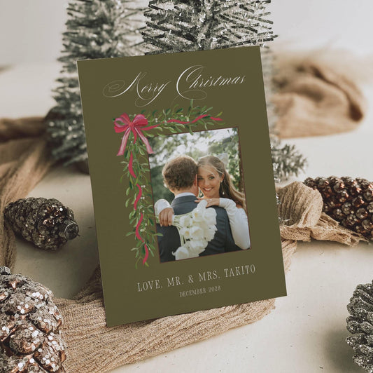 Olive green newlywed Christmas photo card with mistletoe garland and red bow, styled with frosted pinecones and silver trees