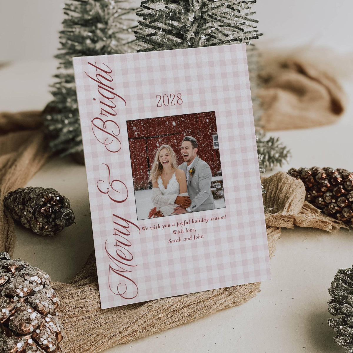 Merry and Bright first Christmas married photo card in red gingham standing among frosted pinecones and silver trees